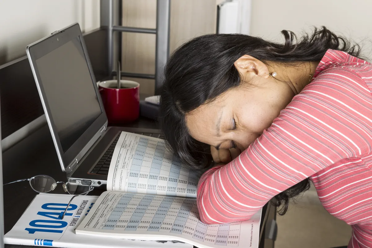 Mature Asian woman with head down on income tax tables booklet with computer, coffee  cup and glasses on desk