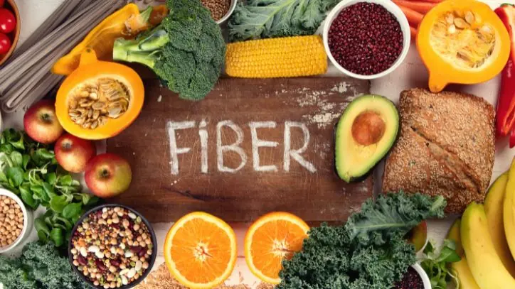 A wooden cutting board with the word "fiber" written on it, surrounded by fiber rich foods